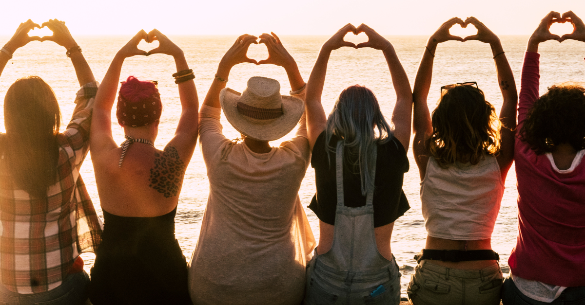 Five women sit on a beach looking at the ocean with their hands in the air forming love hearts